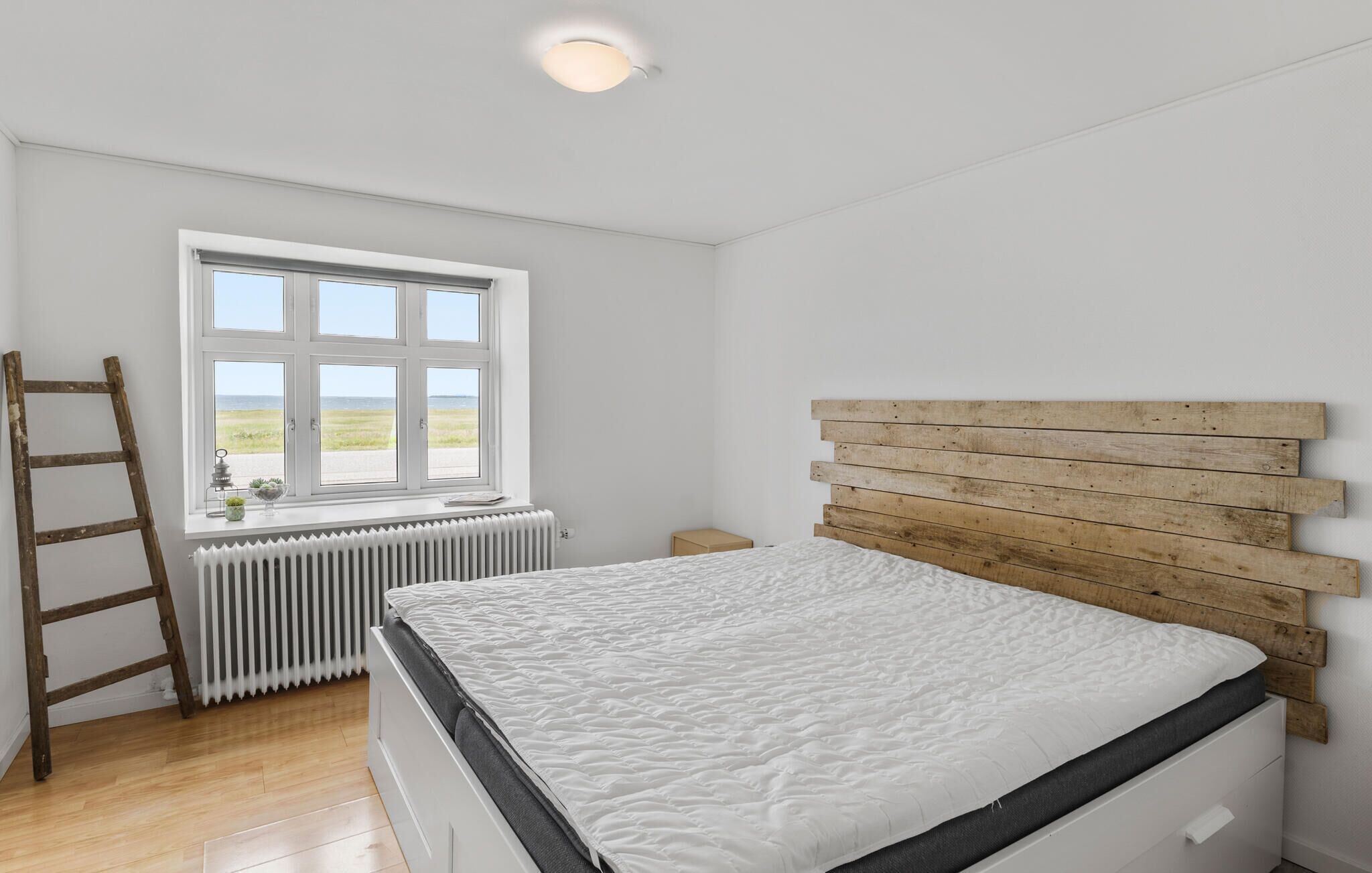 Une chambre avec une tête de lit en bois, une fenêtre donnant sur la mer, un radiateur et une échelle en bois.