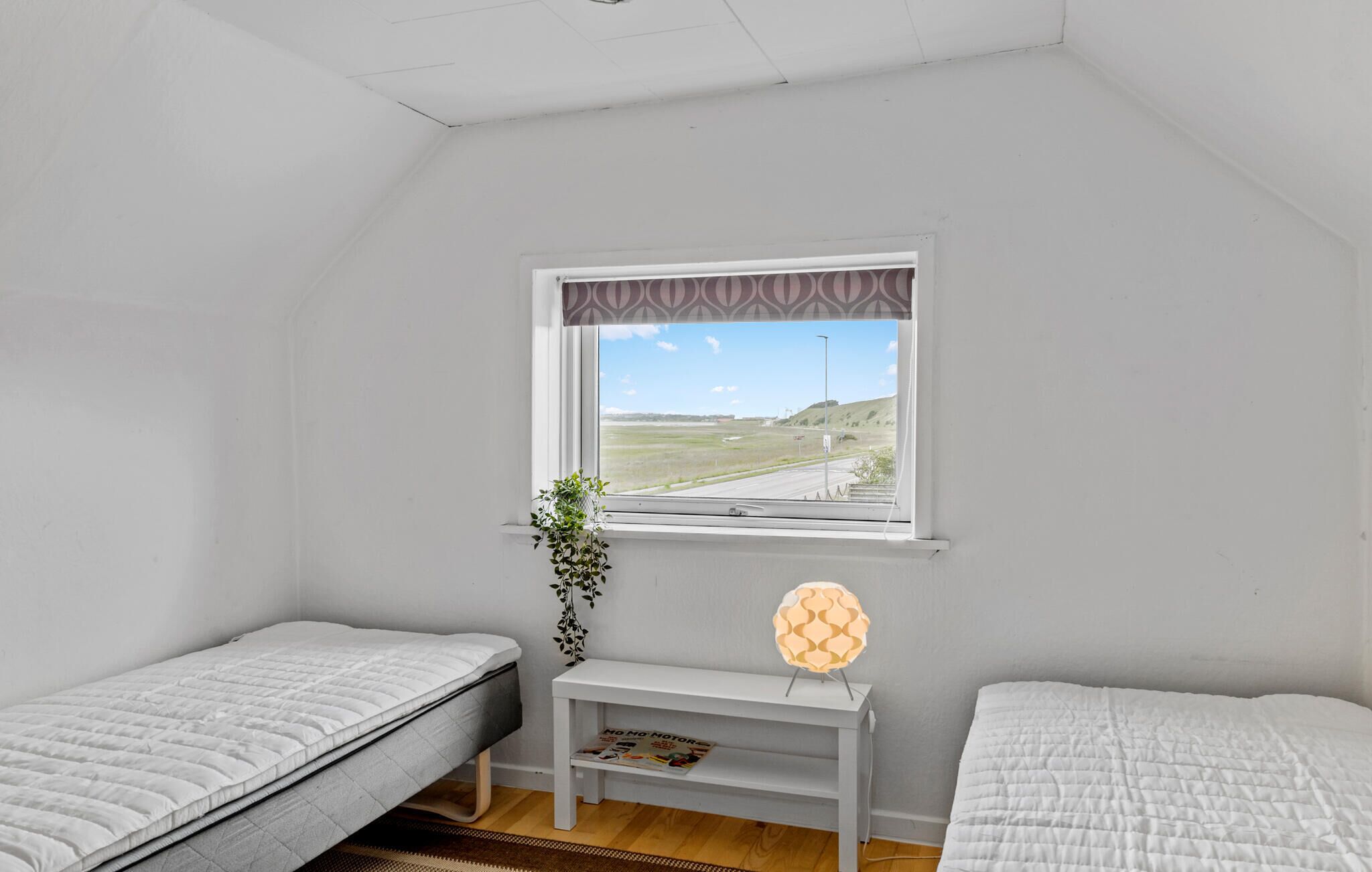 Una habitación pequeña con dos camas, una ventana que da a la vista de una carretera y colinas, una mesita con una revista y una planta.