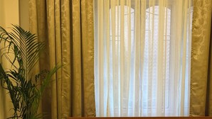 Habitación de hotel con cama, dos almohadas, mesita de noche, planta en maceta y ventana con cortinas.