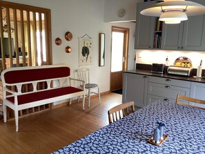 A room with a wooden floor, a white sofa with red cushions, a dining table with a blue floral tablecloth, and a kitchen area with grey cabinets and a hanging light fixture. - 4 Star Holiday Home in Heberg (Heberg)