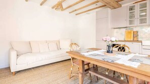 A cozy living space with a wooden dining table, a white sofa, and a kitchen area with cabinets and a hanging light fixture.