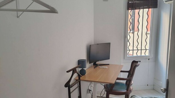 A small room with a wooden desk, a chair, a television, and a window with a metal grill.