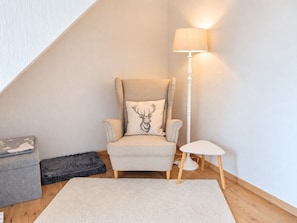 Une pièce d’angle avec un plafond haut, un fauteuil beige orné d’un coussin à motif de cerf, une petite table blanche, un lampadaire, un canapé gris et un tapis blanc.