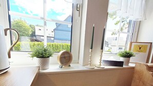 A window with a view of a residential area, a wooden clock, and two potted plants on a windowsill. - FeWo Merle Altastenberg – Nature & Active Holidays (Winterberg)