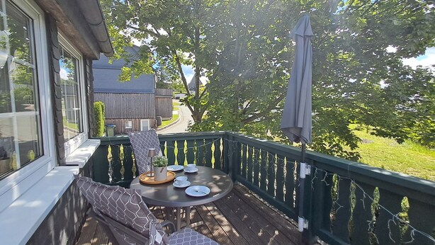 A covered outdoor seating area with a table set for two, a bench, and a view of a rural street. - FeWo Merle Altastenberg – Nature & Active Holidays (Winterberg)