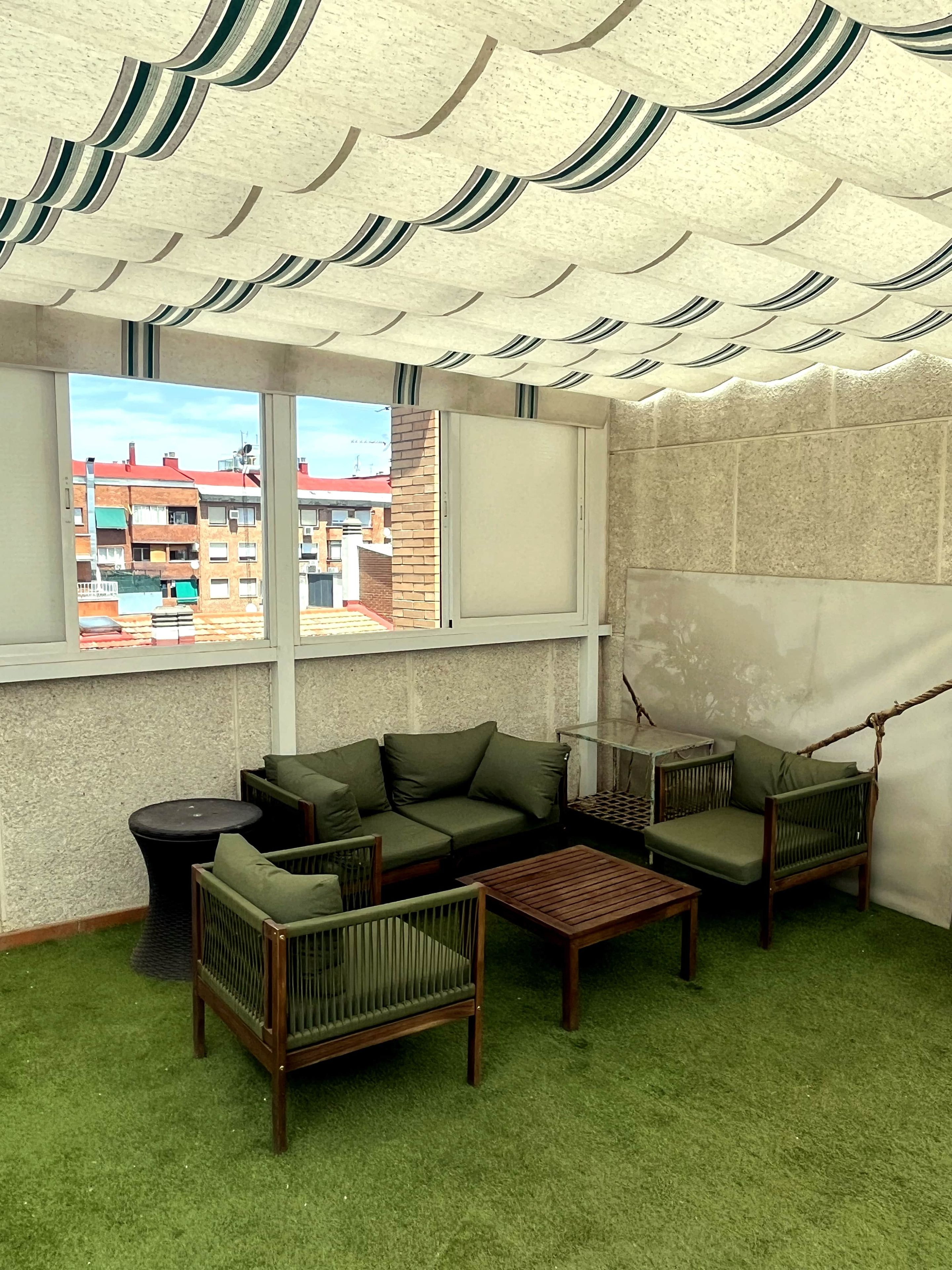 A room with a green sofa, a wooden coffee table, and a hanging chair. There are windows with a view of buildings outside.
