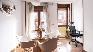 Una habitación moderna con una mesa de vidrio, sillas blancas, una silla de oficina negra, un espejo redondo y vistas a un edificio a través de una ventana.