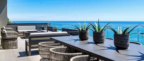 An outdoor seating area with wicker furniture and potted plants, overlooking a clear blue sea.