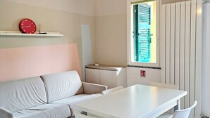 A modern living room with a sofa, a dining table with chairs, a wall-mounted air conditioner, a clock, and a window with blinds. - Ligurian Oasis (Pietra Ligure)