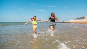 Duas pessoas caminham em águas rasas na praia, com edifícios ao fundo.