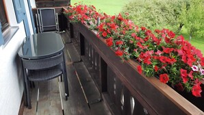 A balcony with a table and chairs, a wooden railing, and a flower box with red flowers. - Vacation rental, 45 square meters, 2nd floor, 1 separate bedroom (Bad Endorf)