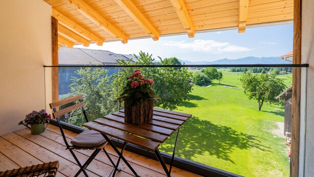 Un balcón de madera con una mesa y sillas, una planta en maceta y vistas a un paisaje verde.