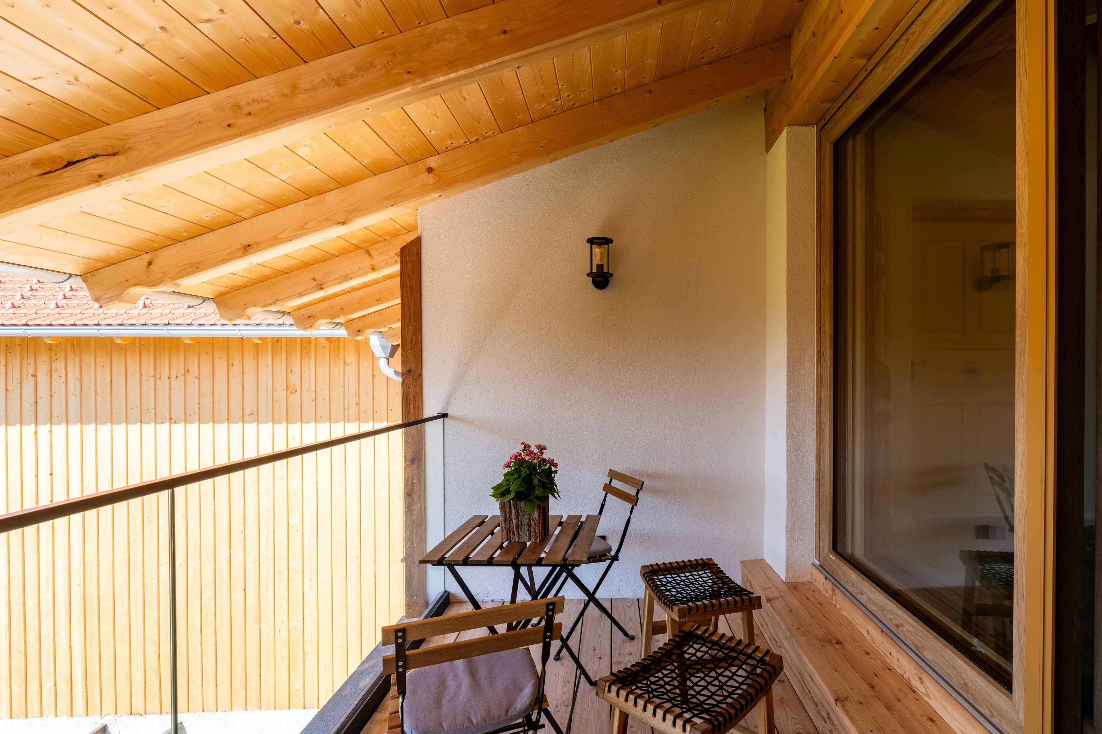 A wooden balcony with a table and chairs, a potted plant, and a large window.