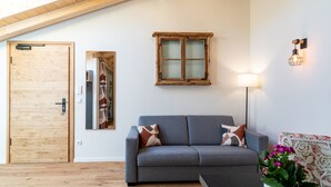 Ein Zimmer mit Holztür, einem grauen Sofa mit gemusterten Kissen, einem kleinen Tisch, einer Topfpflanze, einer Wandleuchte und einem Fenster mit satiniertem Glas.