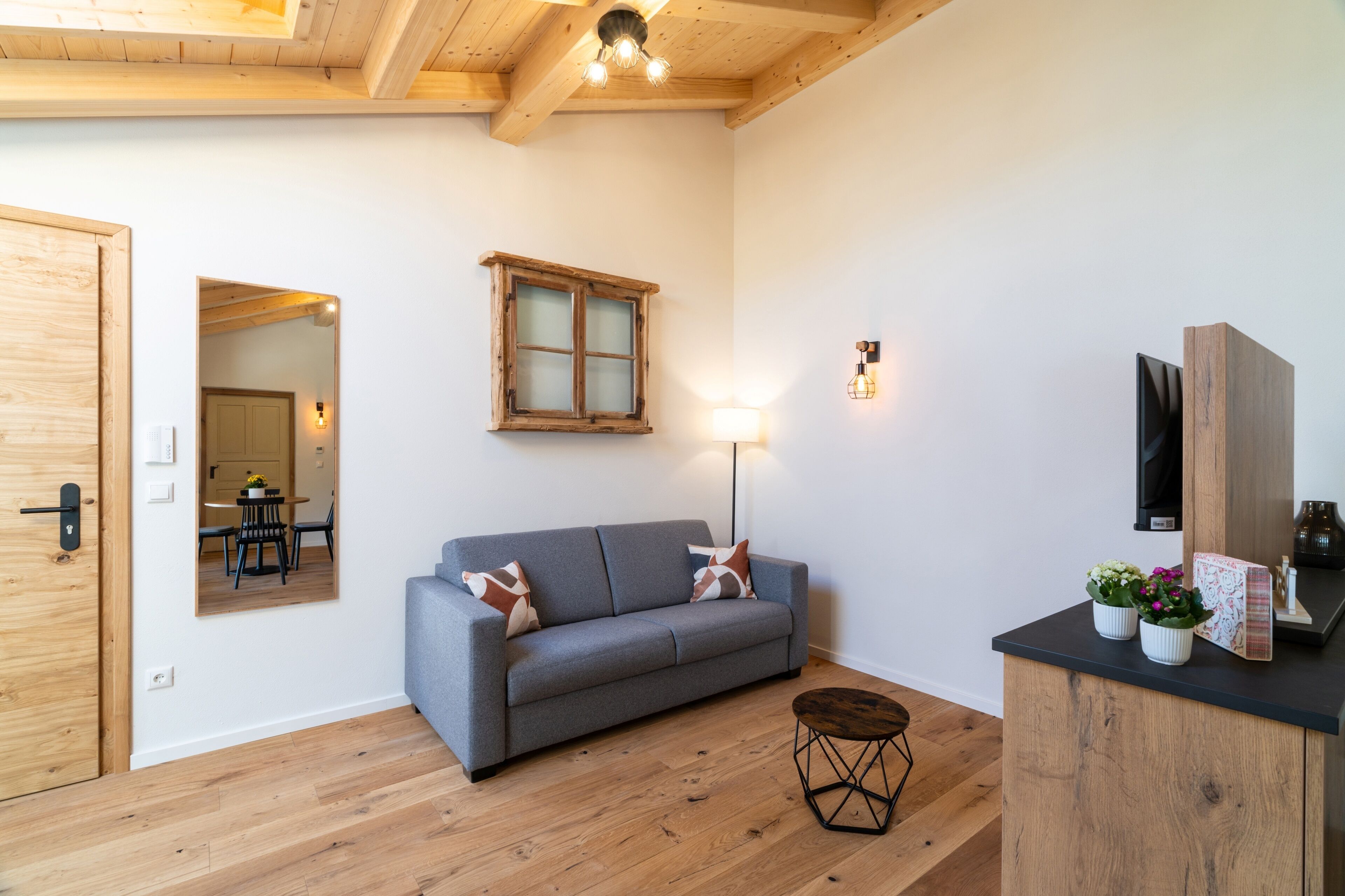 Quarto com sofá cinzento, piso de madeira, janela com moldura de madeira, luminária fixada na parede e uma pequena mesa redonda.