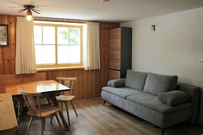 Une pièce comprenant une table à manger en bois, deux chaises, un canapé gris, une fenêtre avec des rideaux et un ventilateur de plafond.