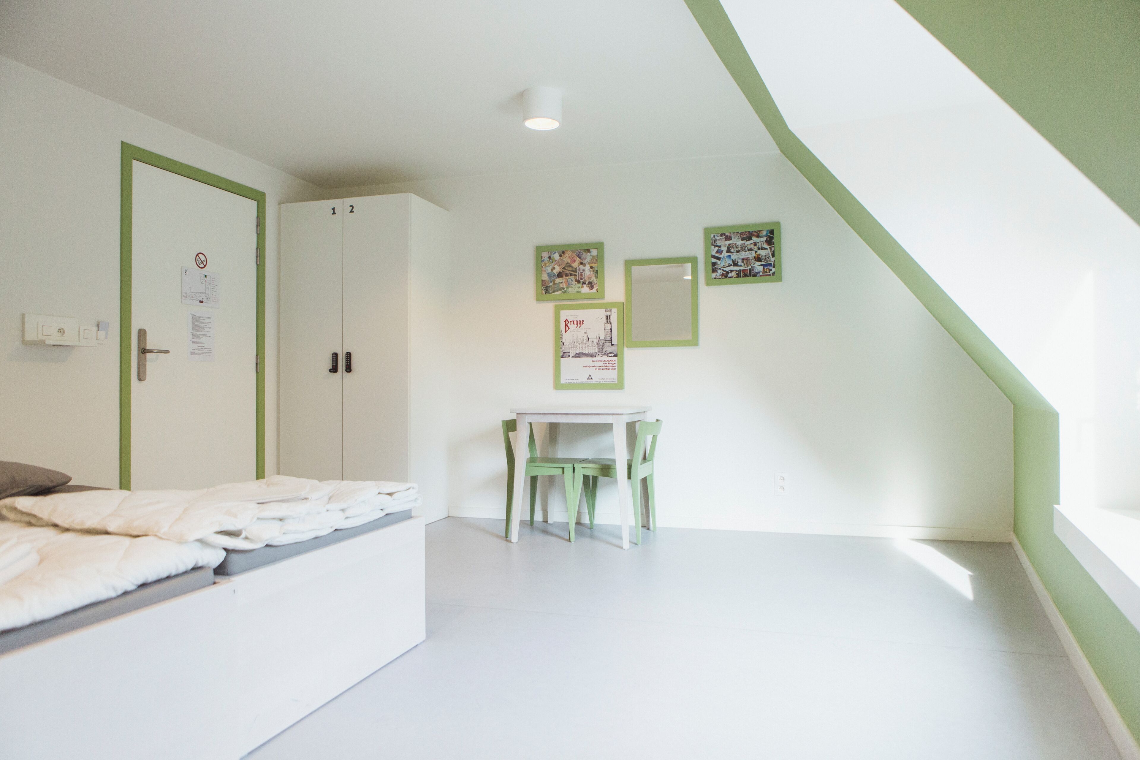 A room with a bed, a desk, and two chairs. There are two doors, one with a green frame and a lock, and another with a white frame and a lock.