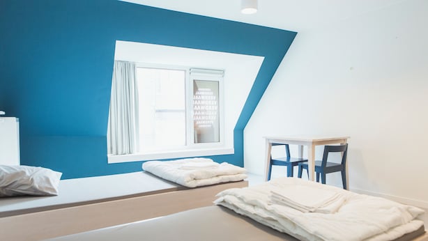 Habitación de hotel con dos camas, una mesita pequeña y un ventanal con cortinas.