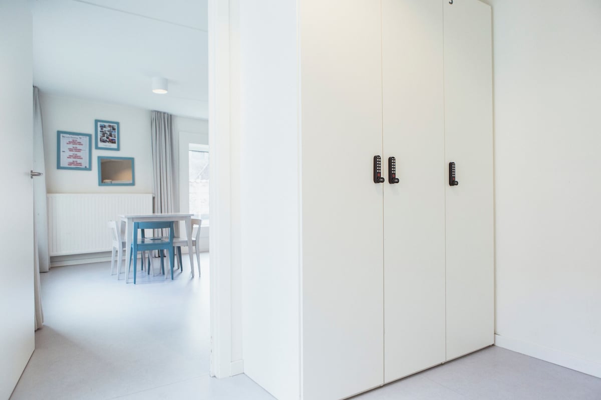 Una habitación con un armario blanco, una mesa con sillas y un ventanal con cortinas.