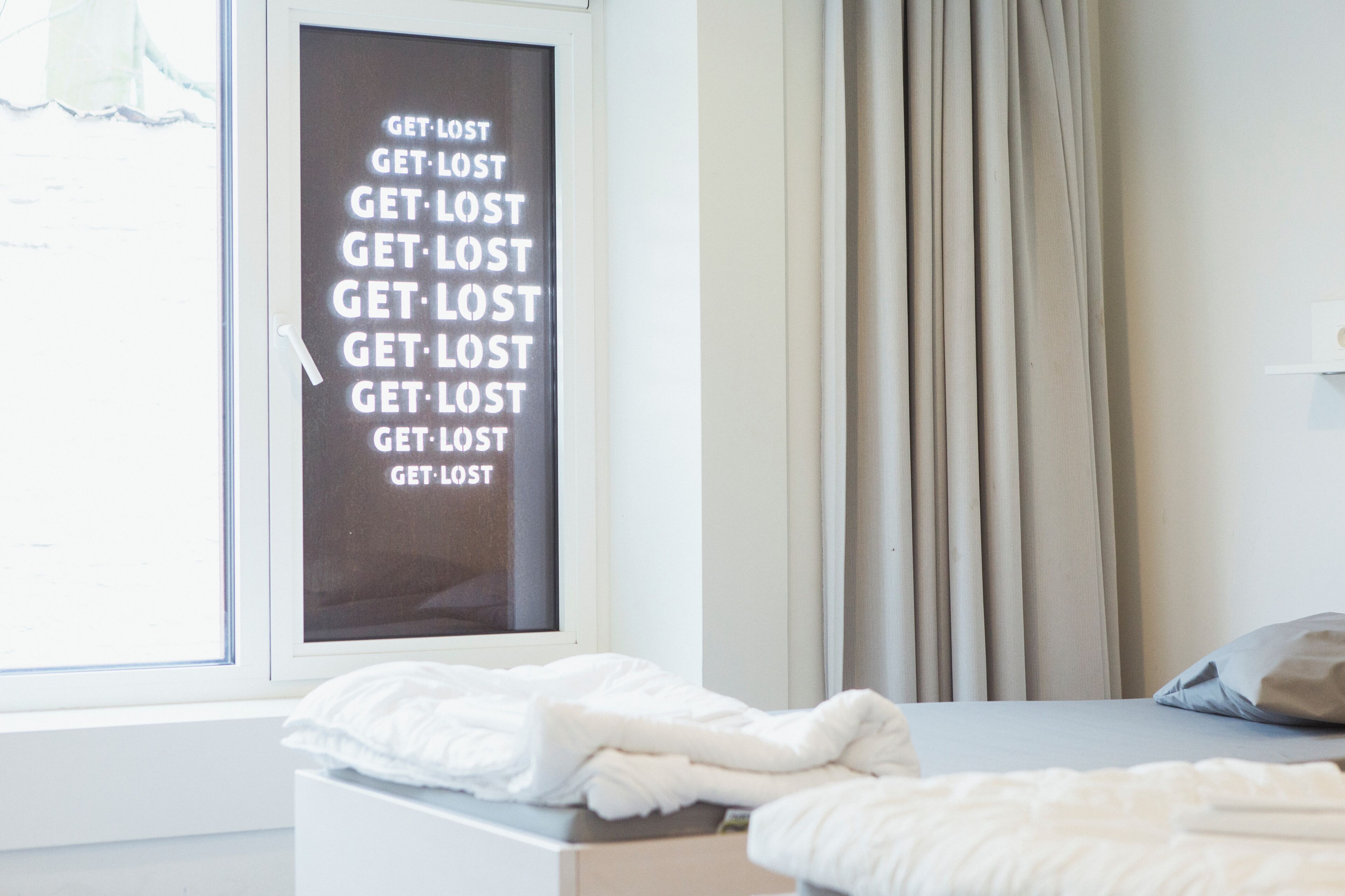 A bedroom with a bed, a window with curtains, and a wall with a repeated text poster.