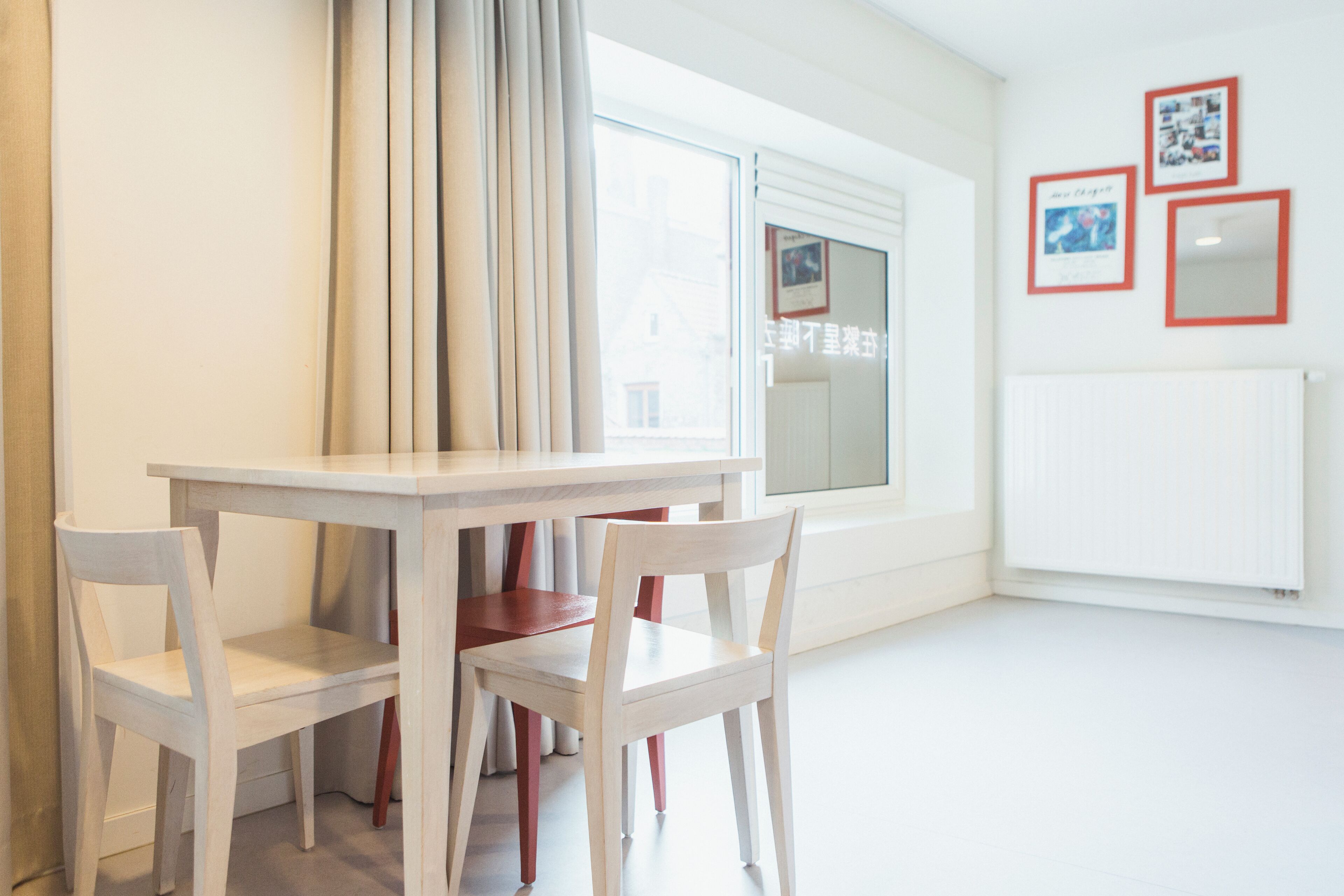 Une pièce avec une table et des chaises blanches, une fenêtre avec des rideaux et un mur orné de tableaux encadrés.