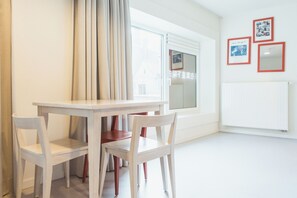 A room with a white table and chairs, a window with curtains, and a wall with framed pictures. - Snuffel Hostel (Bruges)