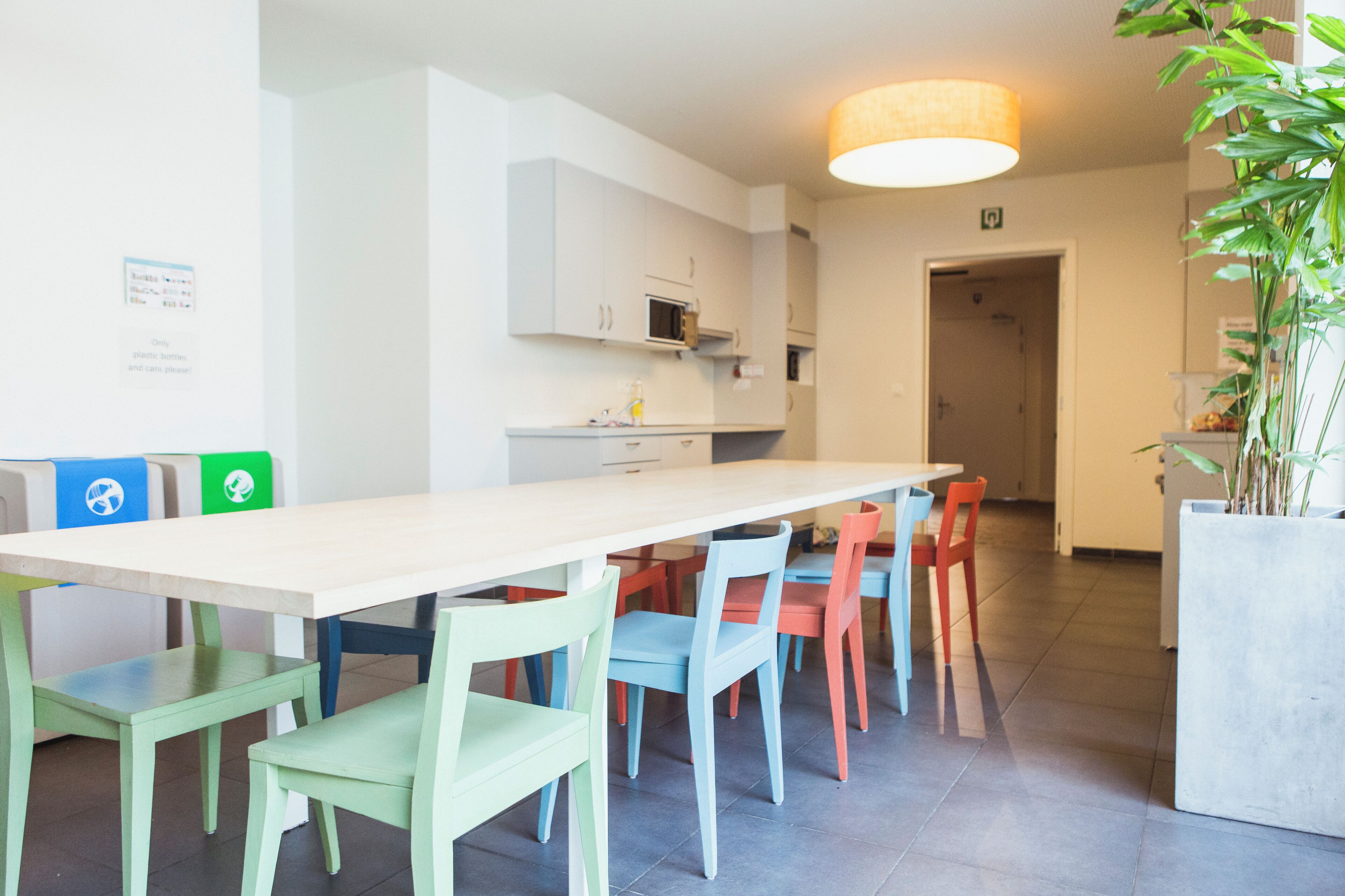 Een moderne keuken met witte kasten, een magnetron en een spoelbak. Er is een eethoek met een lange tafel en kleurrijke stoelen.