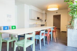 A modern kitchen with white cabinets, a microwave, and a sink. There is a dining area with a long table and colorful chairs. - Snuffel Hostel (Bruges)
