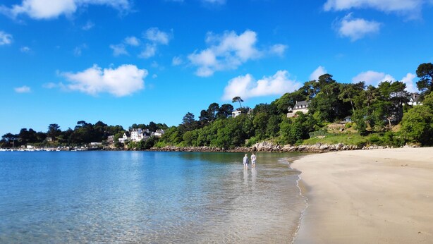 Ein Strand mit klarem Wasser, sandigem Ufer und Häusern im Hintergrund.