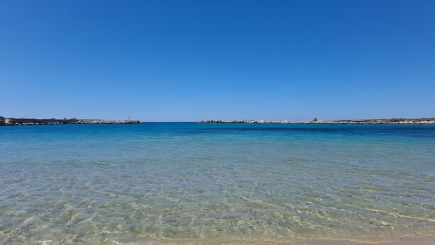 Una playa con agua cristalina, costa rocosa y un muelle a lo lejos.