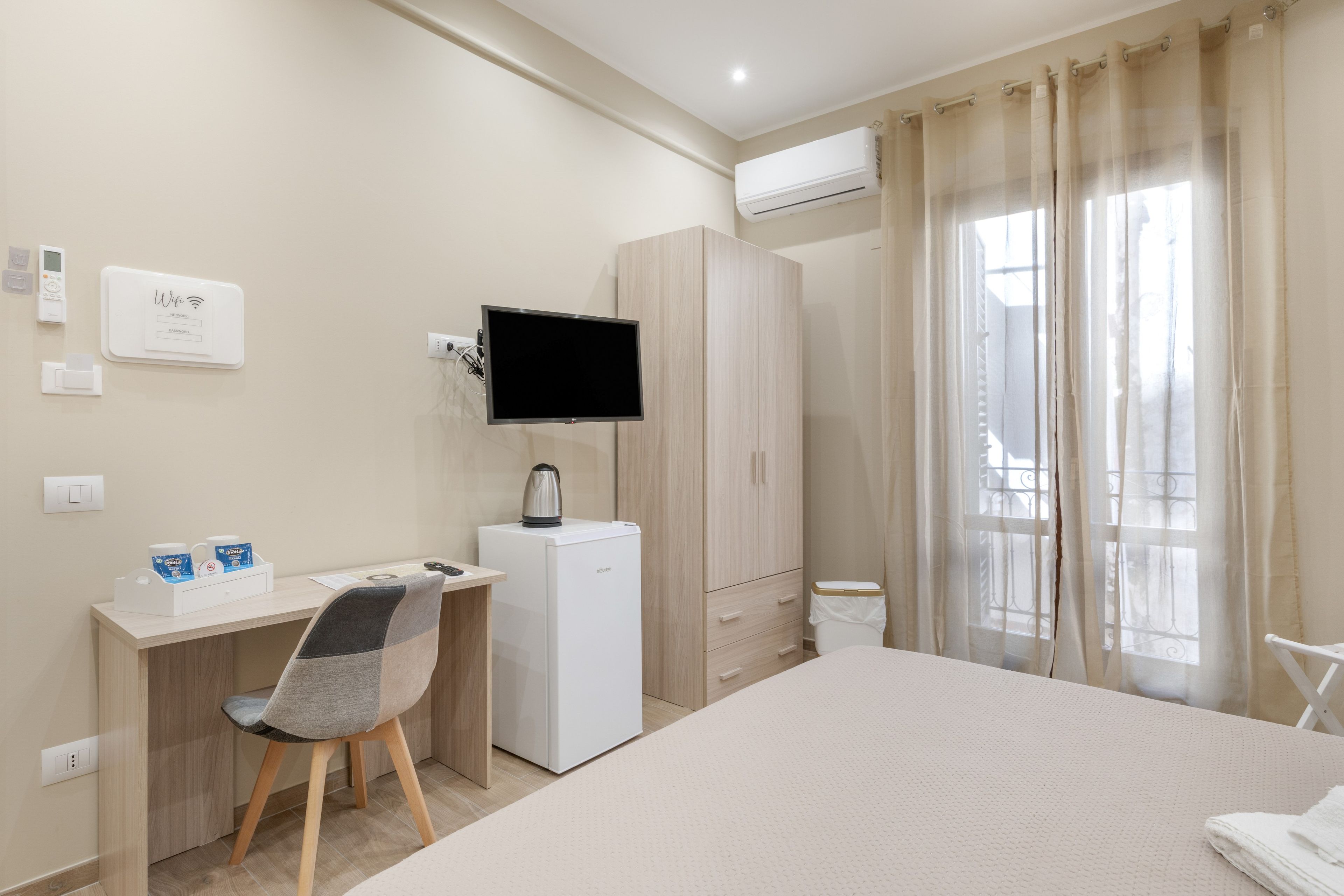 Ein Hotelzimmer mit Bett, Schreibtisch mit Stuhl, Fernseher, Kleiderschrank und einem Fenster mit Vorhängen.