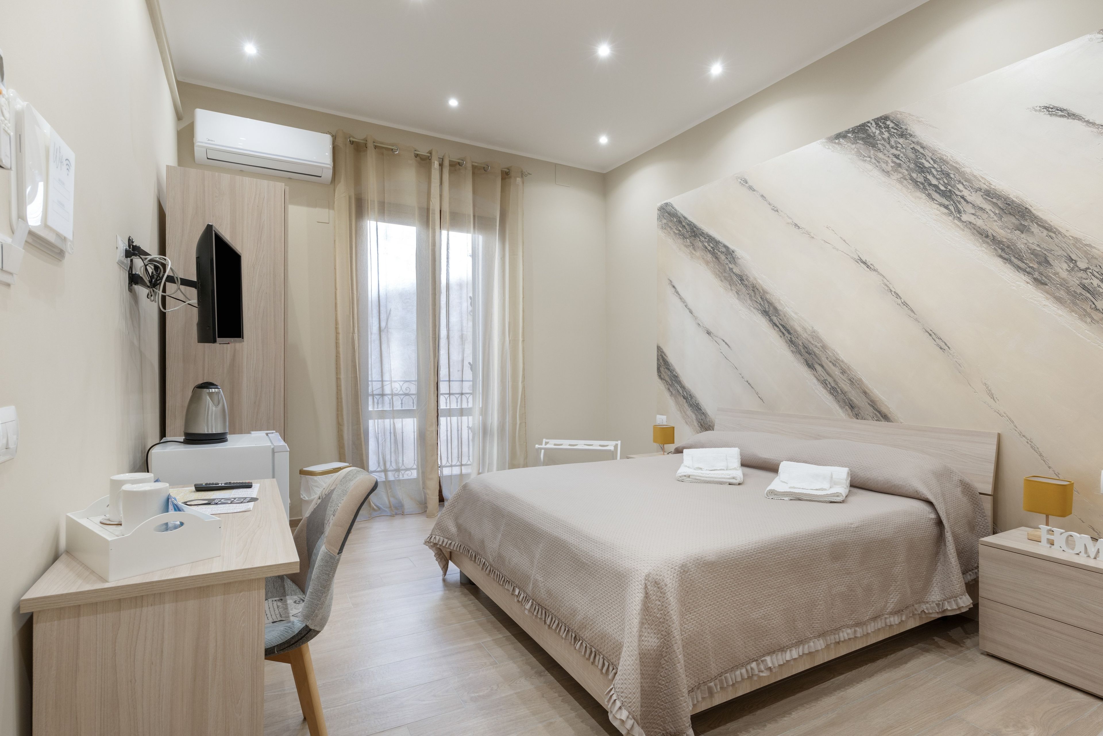 Una camera d'albergo con un letto, una scrivania con una sedia, una macchina del caffè, una televisione e un'ampia finestra con tende.