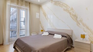 A hotel room with a bed, a nightstand with a lamp, a window with curtains, and a marble wall. - Room 'Stazione' with Balcony, Wi-Fi and Air Conditioning (Palermo)