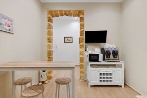 Ein Zimmer mit einem Holztisch, zwei Hockern, einer Kaffeoase mit einer Mikrowelle und einem an der Wand befestigten Fernseher.