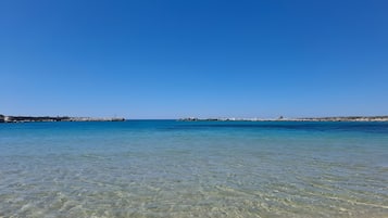 Ein Strand mit klarem Wasser, einer felsigen Küste und einem in der Ferne liegenden Steg.