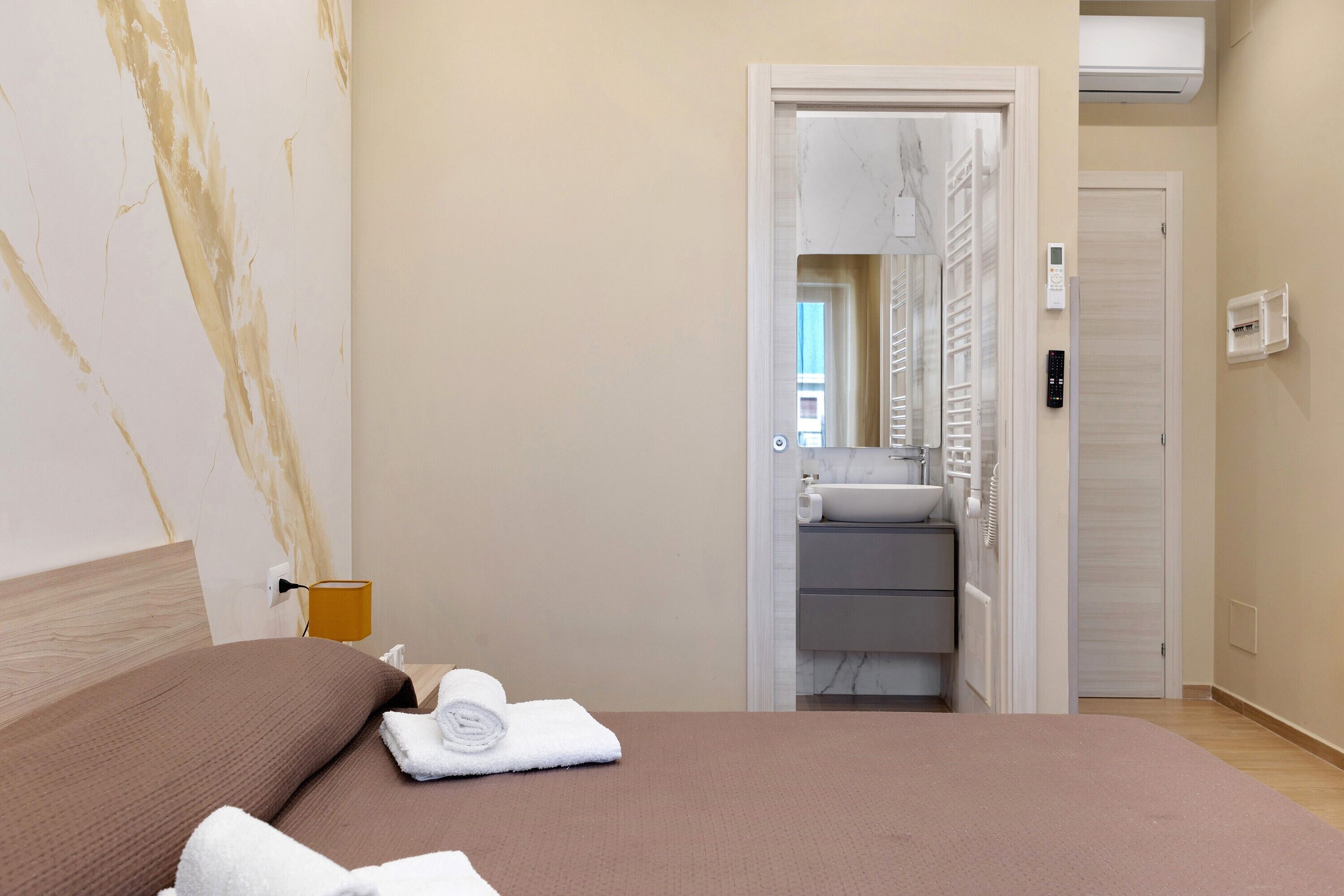 Ein Hotelzimmer mit Bett, einem durchgegangenen Tür erkennbaren Badezimmer und einer Bedienanlage an der Wand.