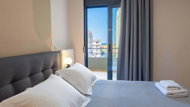 Ett hotellrum med en säng, kuddar, en lampa, en balkong med gardiner och utsikt över byggnader.