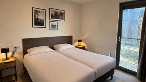 A hotel room with two beds, a small bedside table with a lamp, a large window with a view, and three framed pictures on the wall.