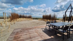 A wooden deck with a table and chairs, overlooking a beach with sand dunes and a clear sky.