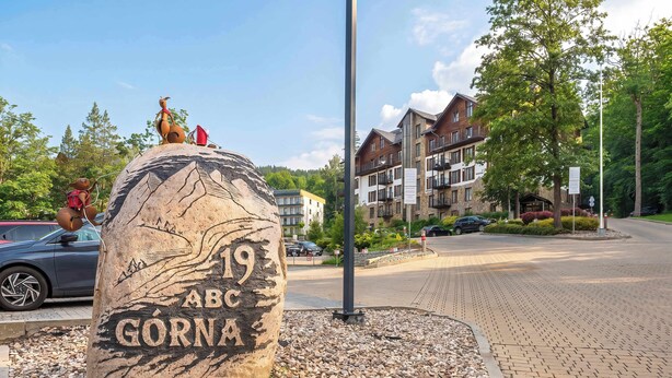 Ein großer Stein mit der eingravierten Aufschrift 'ABC GÖRNIA', umgeben von Kieselsteinen, mit Gebäuden und einer Straße im Hintergrund.