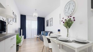Una cocina moderna con armarios blancos, un comedor con una mesa blanca dispuesta para dos personas, y una sala de estar con un sofá y una televisión.