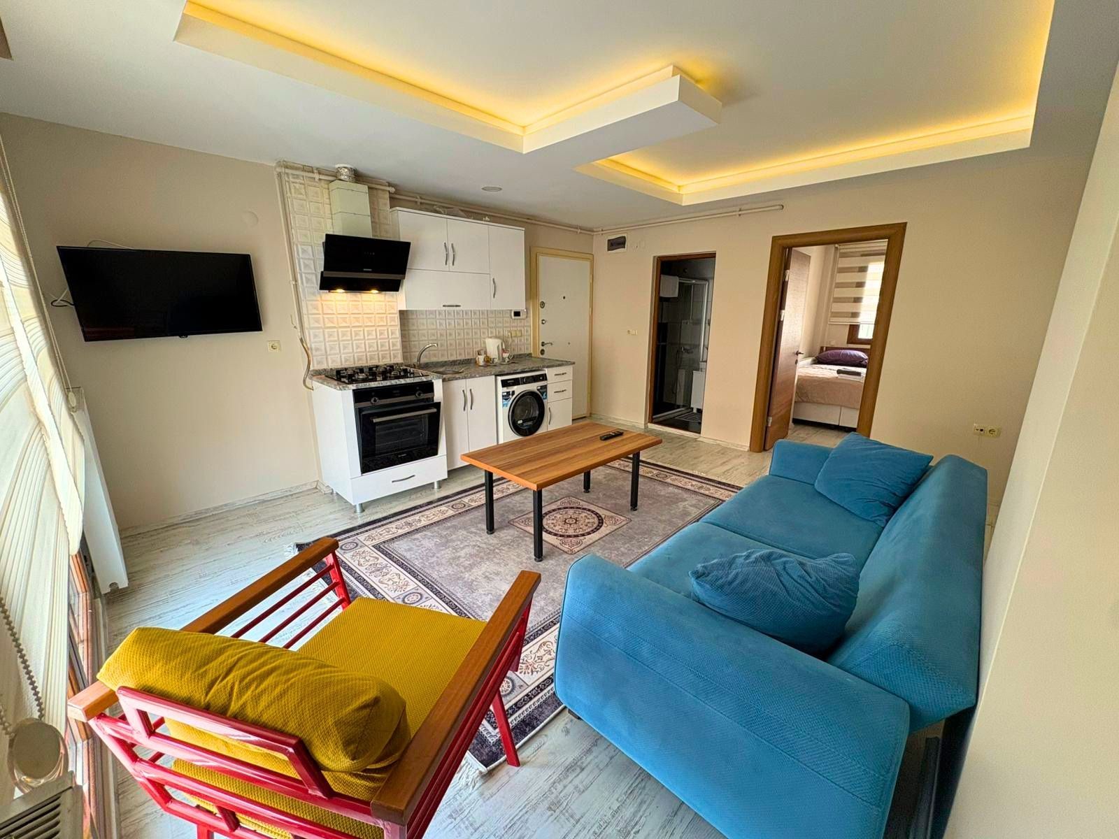 A compact living space with a kitchenette, a sofa, a wooden table, and a chair.