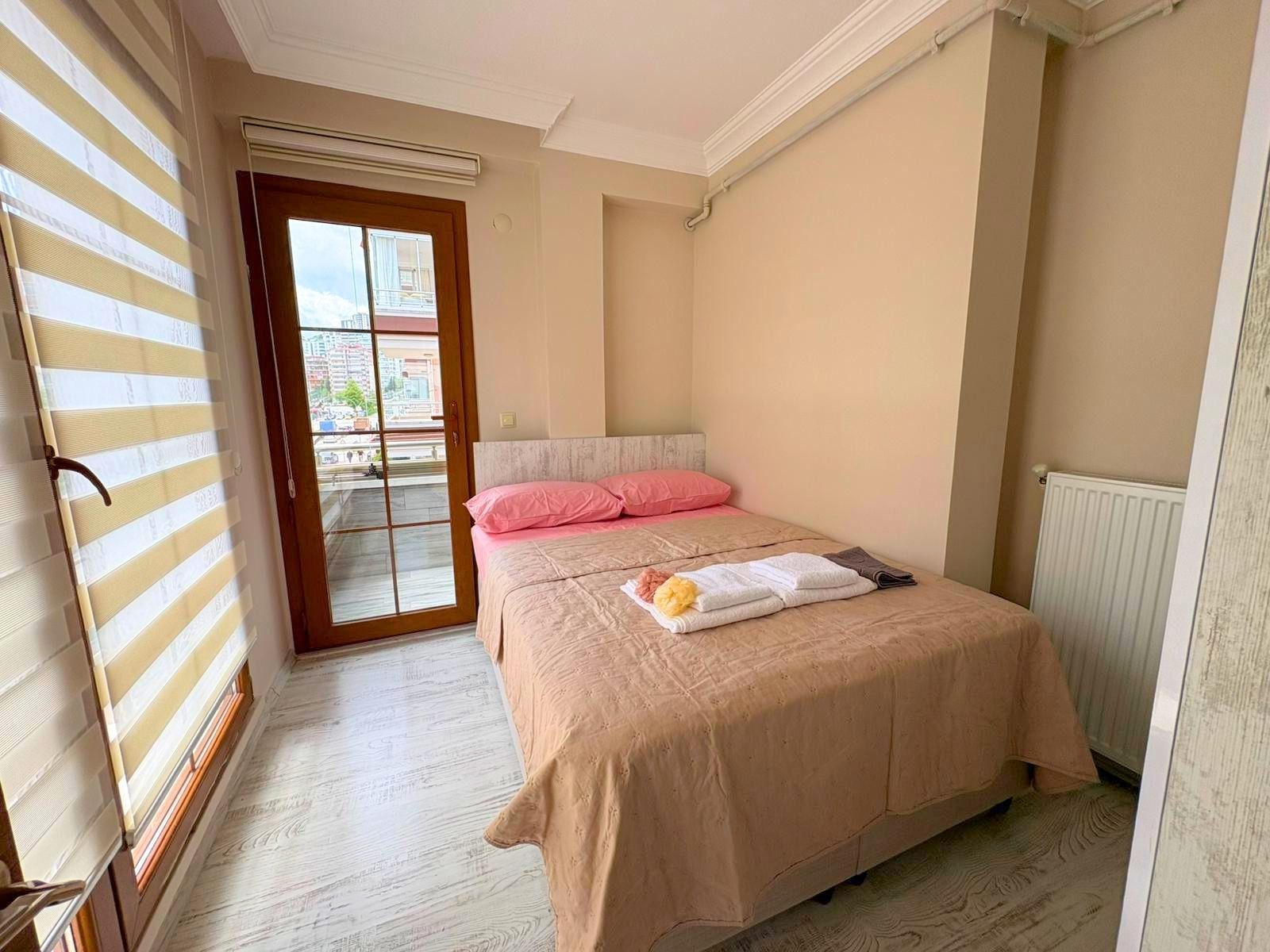 A bedroom with a bed, a window with blinds, a radiator, and a view of buildings outside.