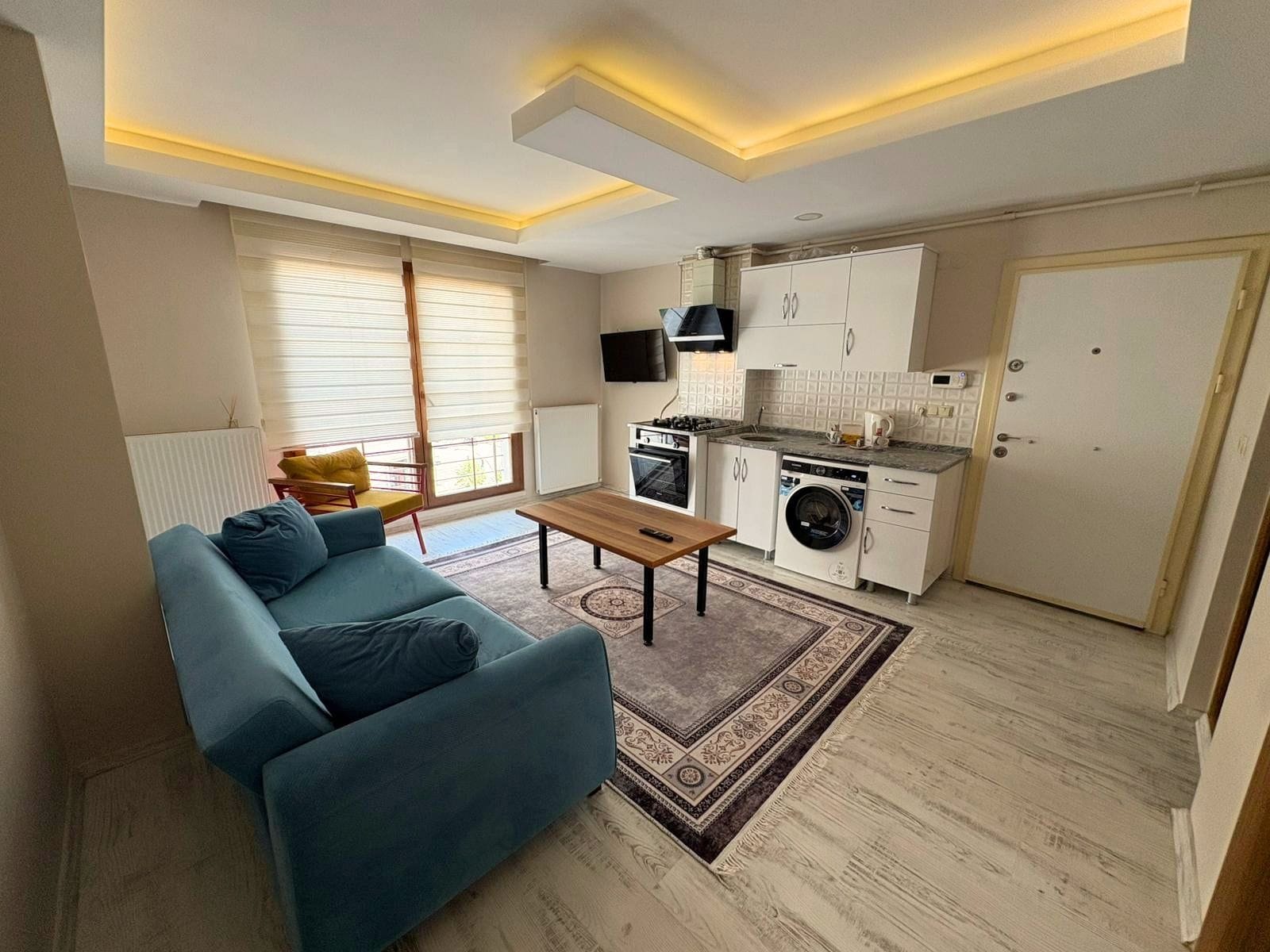 A compact living space with a sofa, a small table, a washing machine, and a kitchenette.