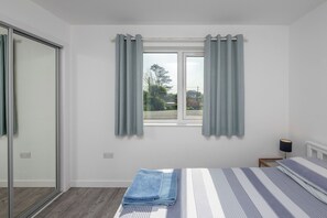 A bedroom with a bed, a window with curtains, a bedside table, and a lamp. - Moranedd Bach (Holyhead)