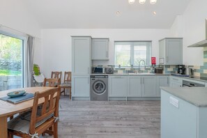 A modern kitchen with a washing machine, a dining area with a table and chairs, and a window with blinds. - Moranedd Bach (Holyhead)