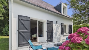 Un bâtiment blanc doté de grandes fenêtres et de volets, une terrasse avec des chaises bleues et, au premier plan, des fleurs roses.