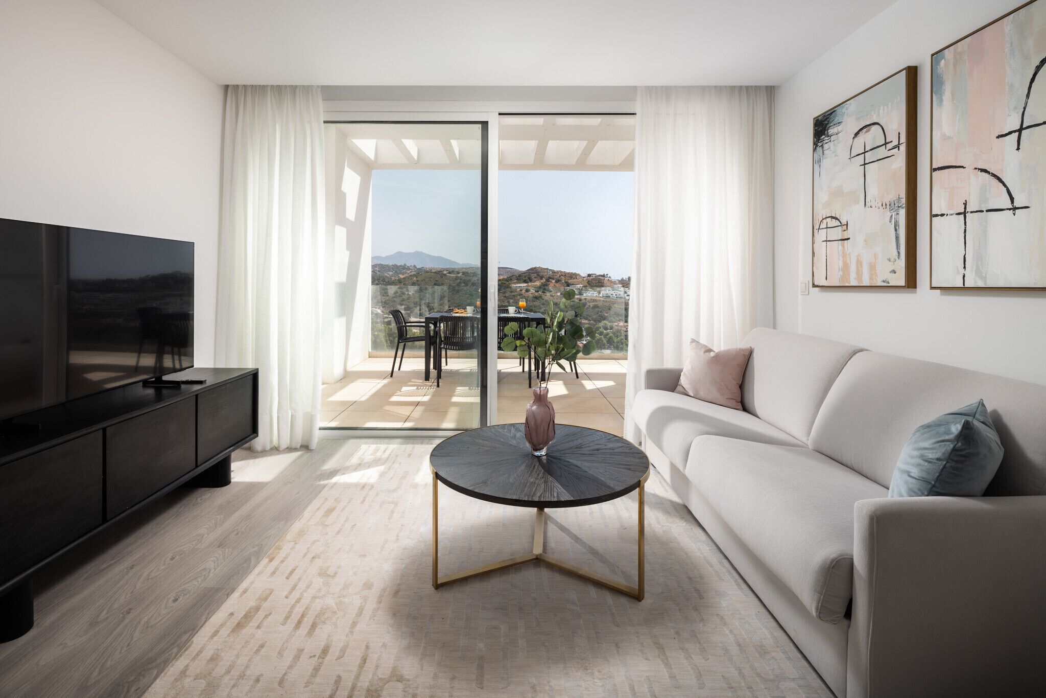 A modern living room with a flat-screen TV, a round coffee table, a sofa, and a balcony with a table and chairs.