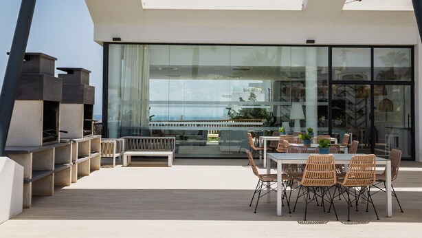 A modern outdoor dining area with a glass wall, wooden furniture, and a view of the sea. - Casa Banderas Sea & Valley Views by Alfresco Stays (Mijas)