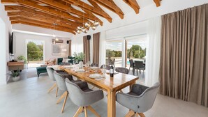 Un amplio comedor con una mesa de madera y sillas tapizadas grises, una cocina moderna con vigas de madera y un gran ventanal con vistas a una piscina.